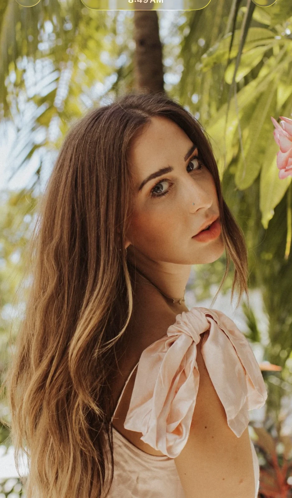 Elizabeth Faye under palm trees, hair down, a pink bow on her shoulder